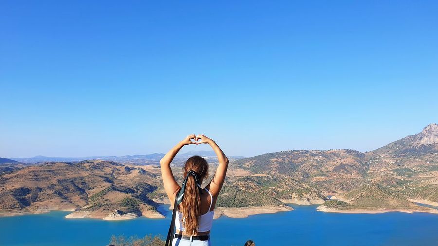 Rear view of woman making heart shape against mountain and sky