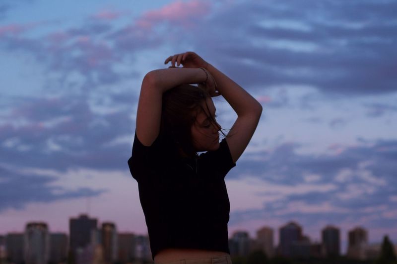 Young woman with arms raised standing in city against sky during sunset