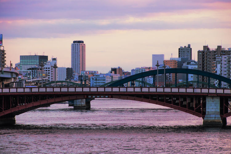 Bridge over river by buildings against sky during sunset