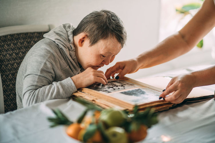 Girl points a smiling elderly woman with down syndrome to a photo in an old album.