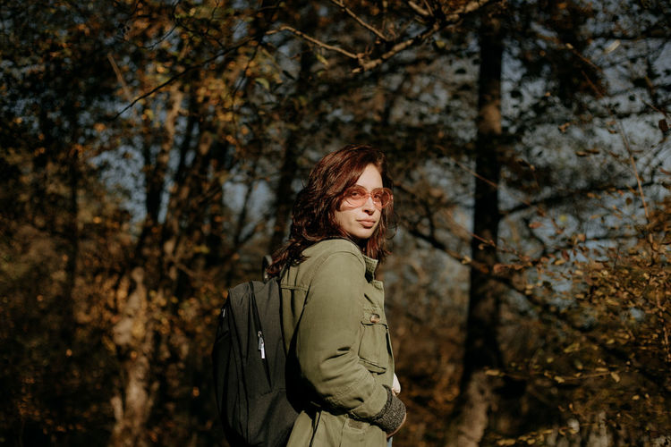 Young woman looking away while standing next to a tree in forest during autumn 