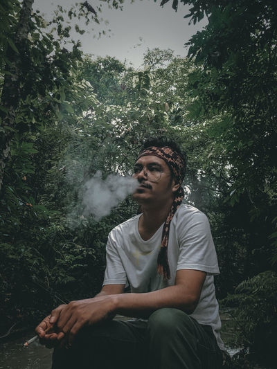 Young man smoking cigarette while sitting in forest