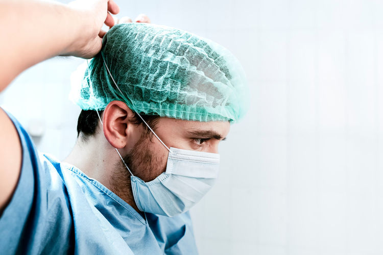 Side view of serious professional male surgeon in uniform and cap putting on medical mask while preparing for operation in hospital