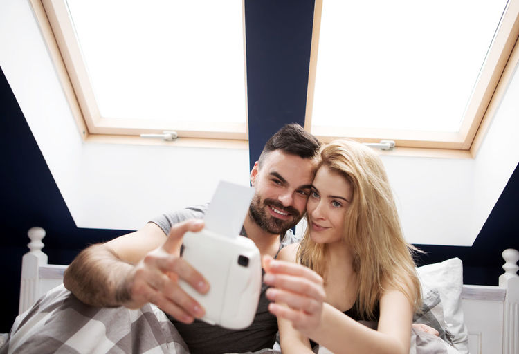 Young man with woman taking selfie from instant camera on bed at home
