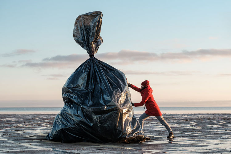 A woman pushes a huge black trash bag on the shore