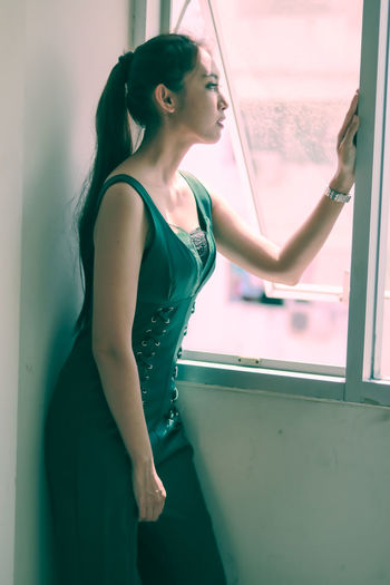 Young woman looking away while standing against window