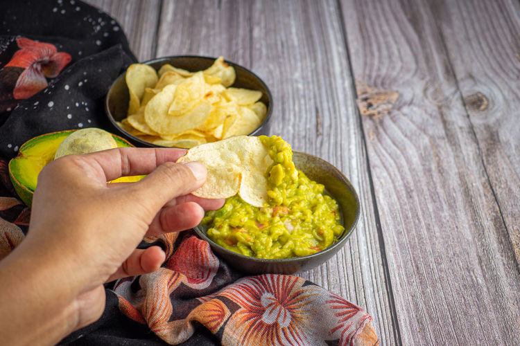 Fresh guacamole with potato chips in bowl and fresh avocados in basket on wooden table. 