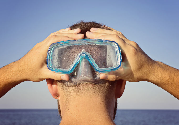 Rear view of man holding swimming goggles at beach against sky