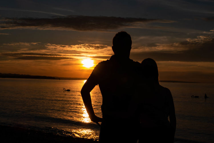 Silhouette pair standing on beach against sky during sunset