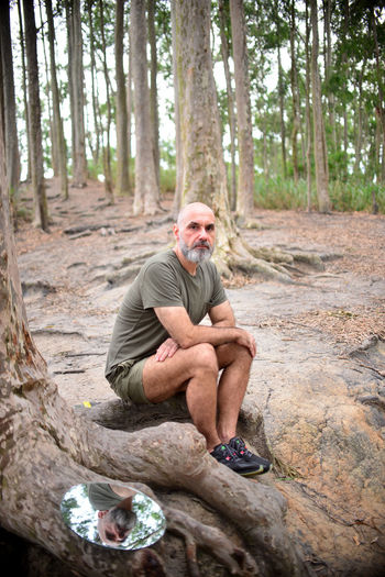 Full length portrait of man sitting by tree trunk in forest