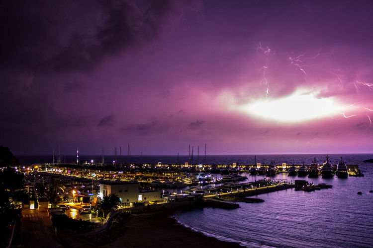Illuminated city by sea against sky at night