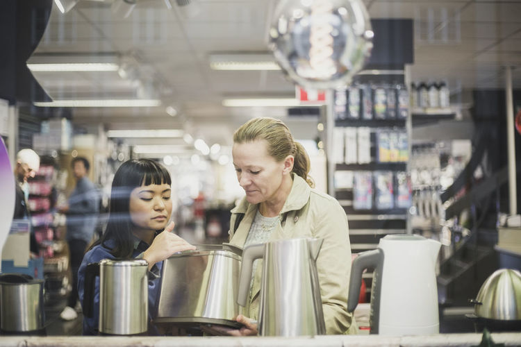 Saleswoman showing kettle to female customer in store seen through glass window