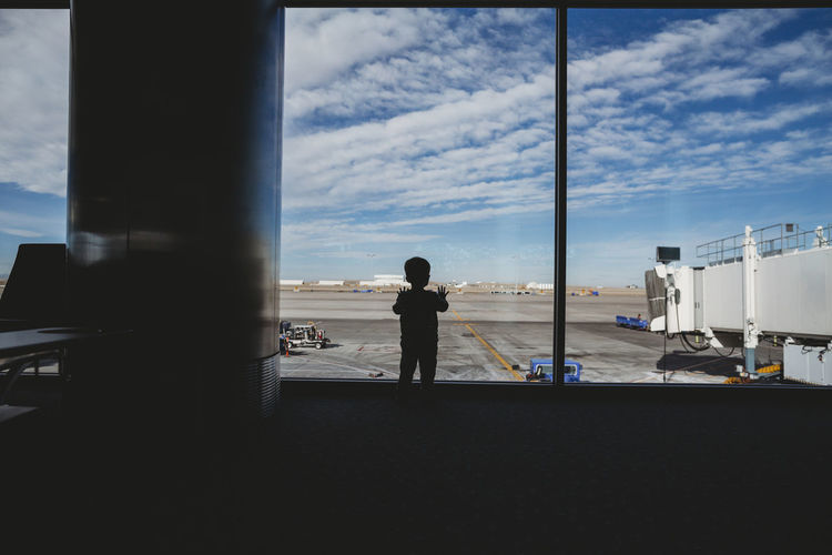 Rear view of silhouette baby boy looking through window at airport departure area