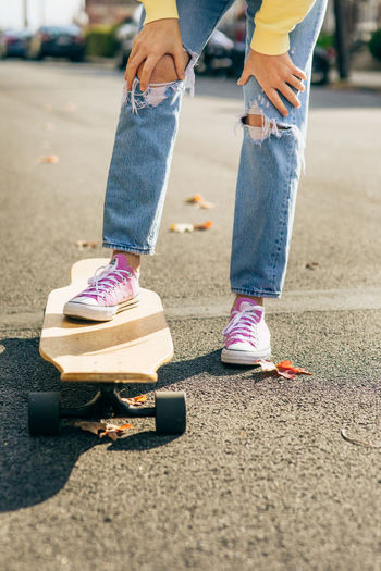 Girl with hands on knees, with one foot on longboard resting on the street in the sun.