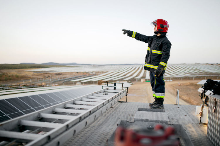 Full body side view of male firefighter wearing uniform and gloves standing on metal roof of fire engine near ladder with hand pointing forward at solar panels in rural farm