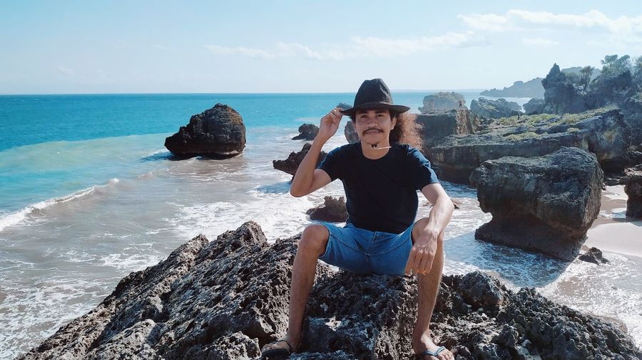 Portrait of man wearing hat sitting on rock against sea