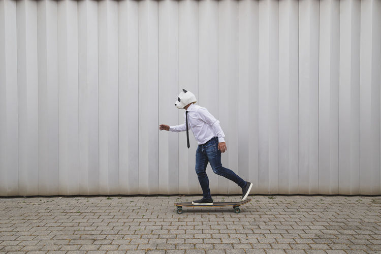 Businessman skateboarding on longboard with a panda bear mask against gray wall person