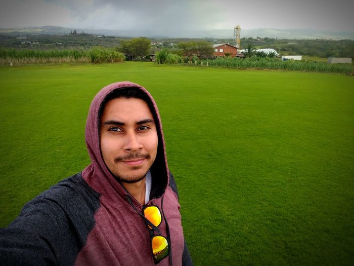 Portrait of smiling young man wearing hood on grassy field against sky