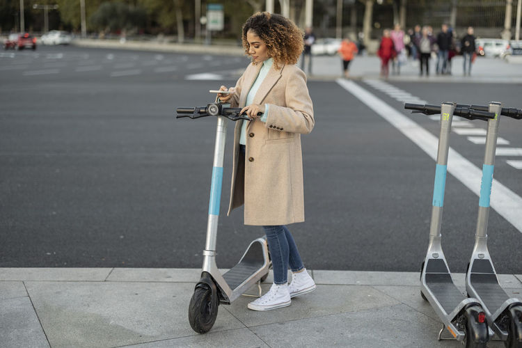 Young woman renting electric push scooter through smart phone on footpath