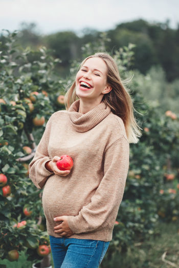 Portrait of pregnant woman holding apple while standing by basket and trees