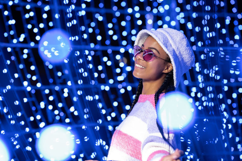 Joyful woman of color with a white beanie braided hairstyle and pink glasses enjoying the christmas 