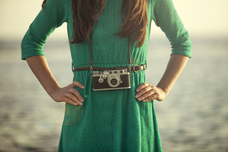 Midsection of young woman with camera standing at beach