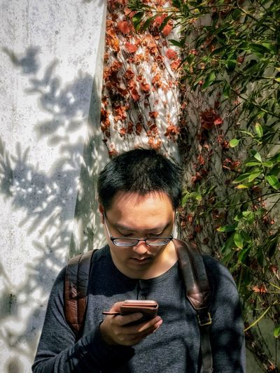 Portrait of young man using mobile phone against ivy covered wall and plants.