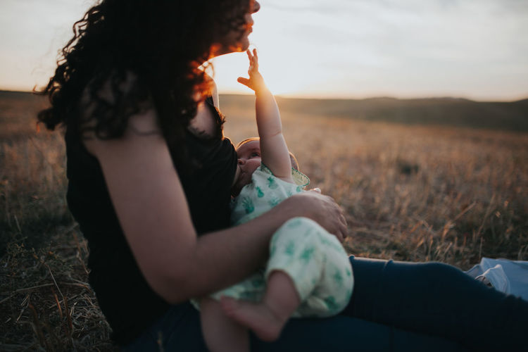 Side view of mother breastfeeding baby while sitting on field against sky during sunset