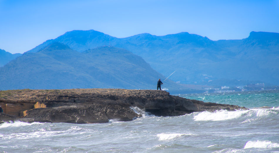Man fishing on rock formation at sea by mountains against sky