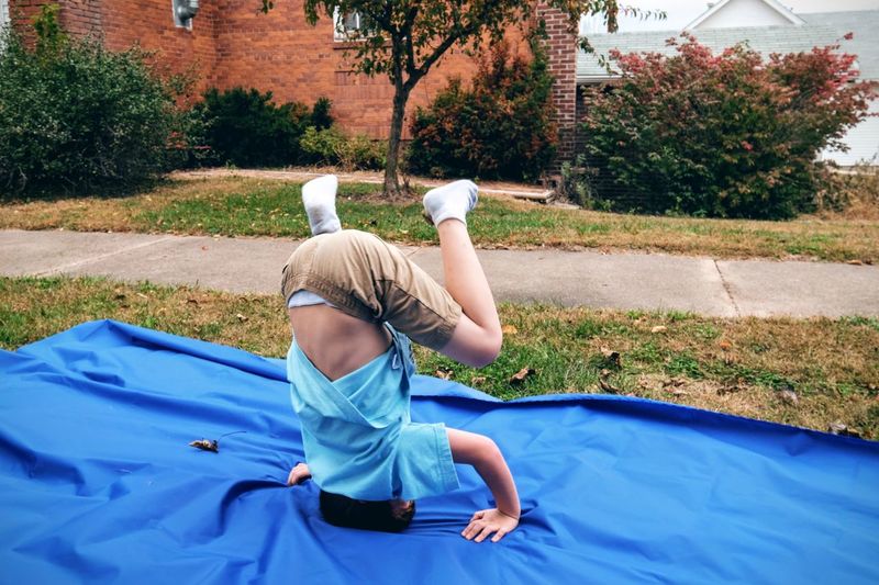 Boy practicing handstand on blue fabric at grassy field against sky