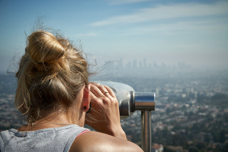Woman looking at cityscape through coin-operated binoculars against sky
