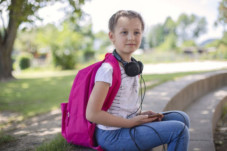 Cute happy girl with a pink backpack sits in a schoolyard or street with headphones around her neck