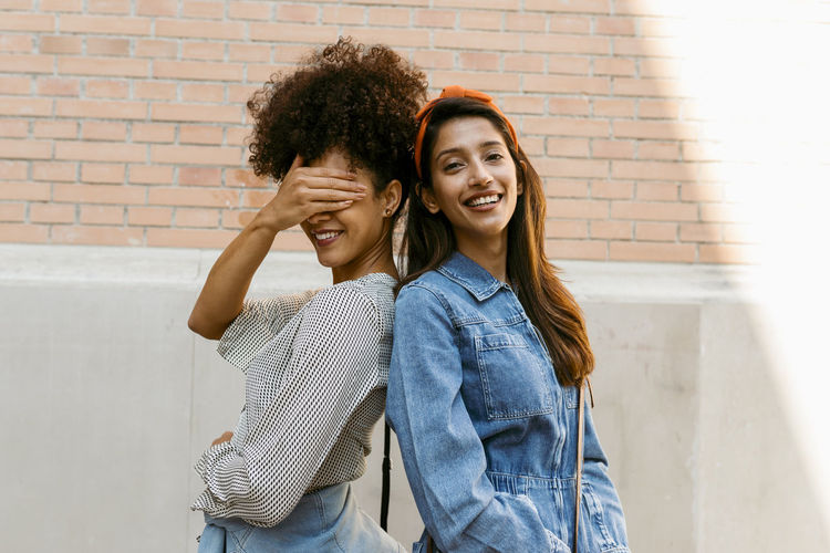 Friend covering eye with hand while standing back to back with woman against wall