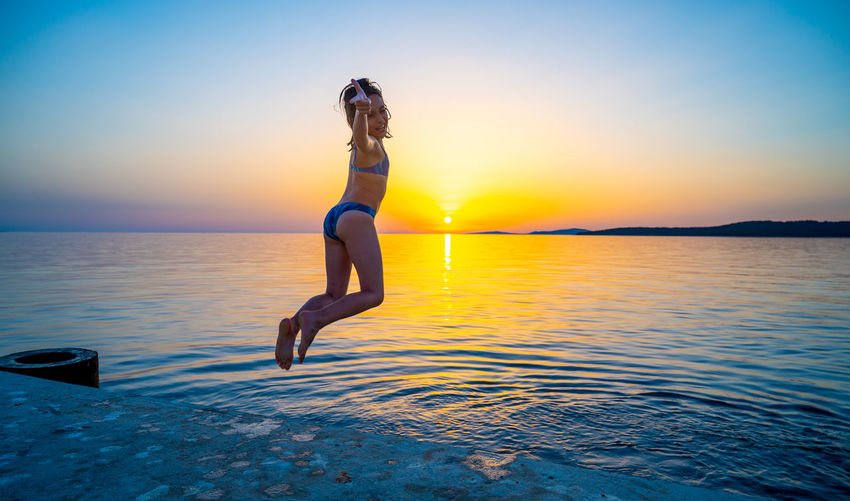 Full length of woman jumping in sea against sky during sunset