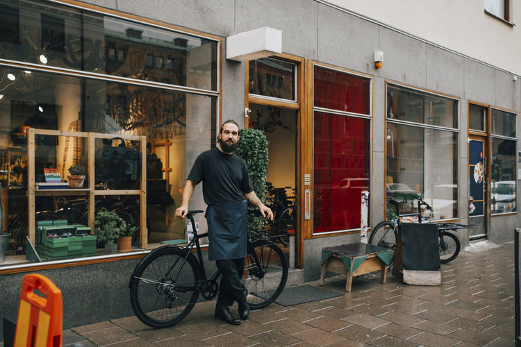 Bearded male owner standing against bicycle outside repair shop