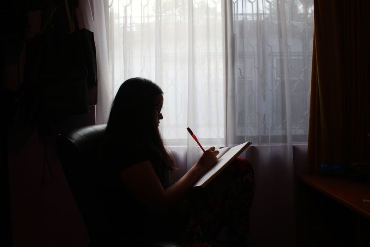 Woman writing in book while sitting on sofa at home