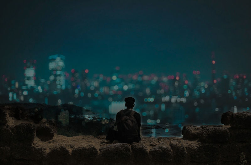 Rear view of man sitting on illuminated cityscape against sky at night