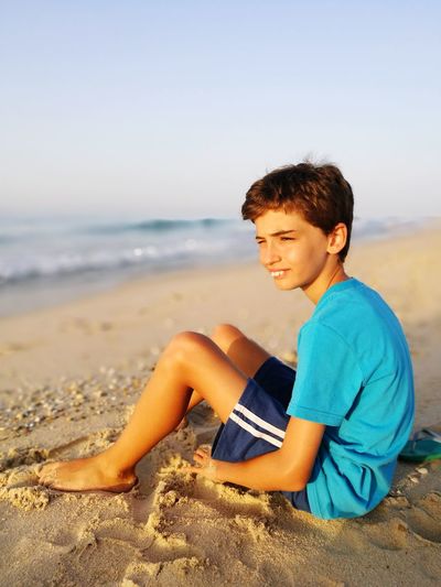 Boy sitting on sand at beach against sky during sunset