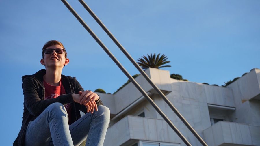 Low angle view of young man wearing sunglasses while sitting on building terrace against clear sky
