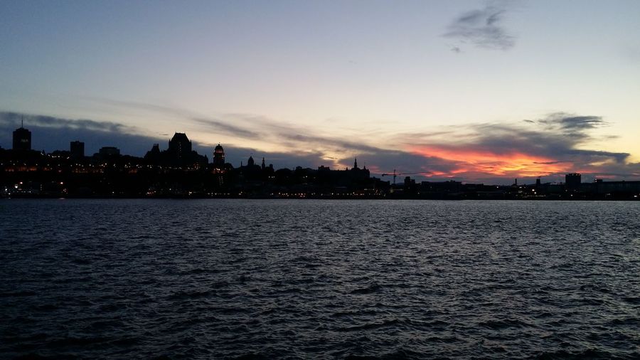Silhouette buildings by sea against sky during sunset