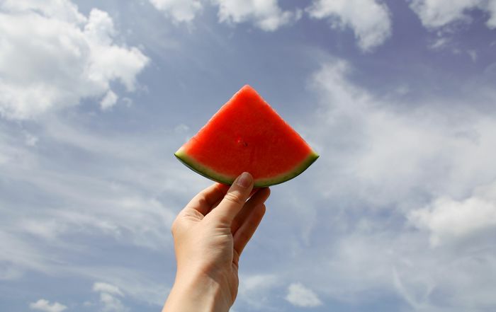 Cropped hand holding watermelon slice against cloudy sky on sunny day