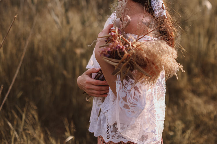 Cropped female figure in a white blouse and long skirt with a bouquet of dried flowers in the field
