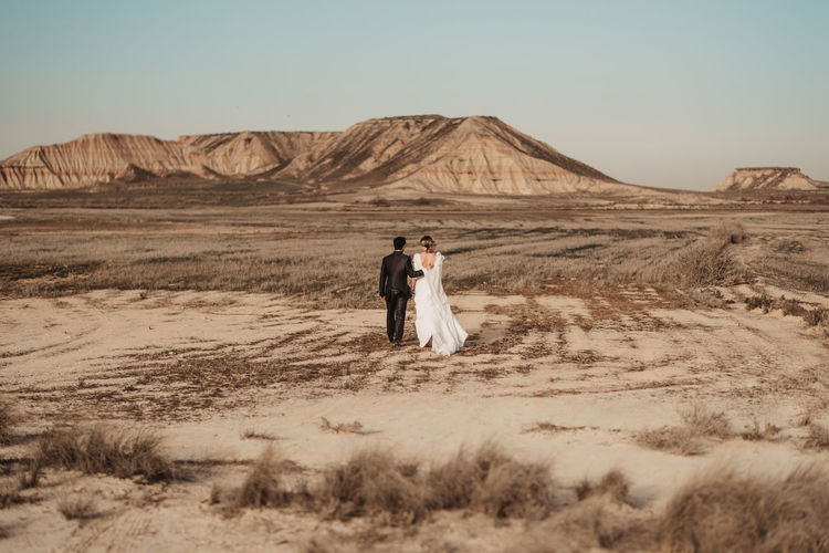 Back view of unrecognizable bride and groom in elegant clothes walking on arid plain towards mountains during wedding celebration in bardenas reales natural park in navarra, spain