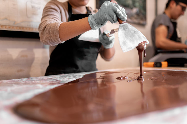 Anonymous employee in gloves using metal spatula to spread liquid chocolate on table while making dessert in confectionery