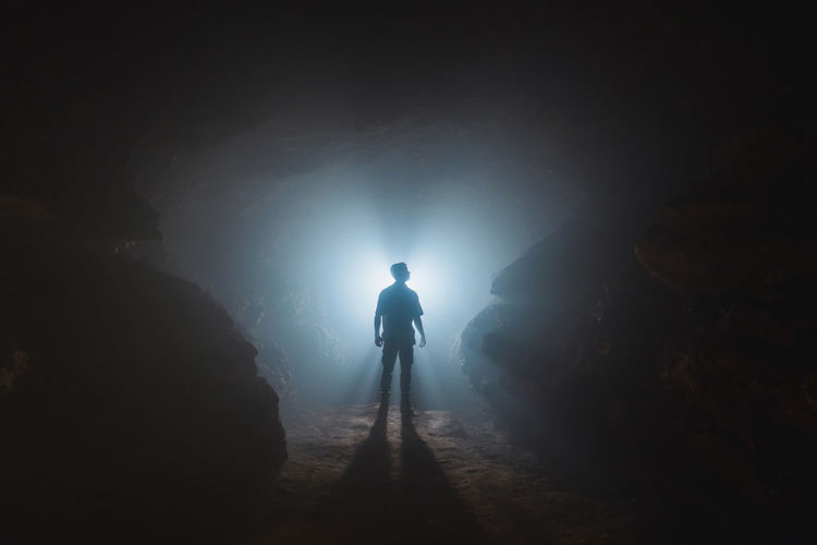 Silhouette of anonymous male explorer standing alone in dark narrow rocky cave against light glowing from entrance