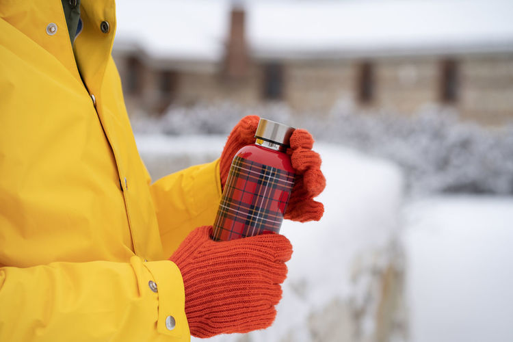 Man hands in orange gloves opening thermos bottle with red checkered pattern in winter outdoors