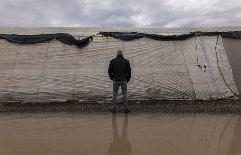 Rear view of adult man in front of plastic greenhouses against cloudy sky in almeria, spain