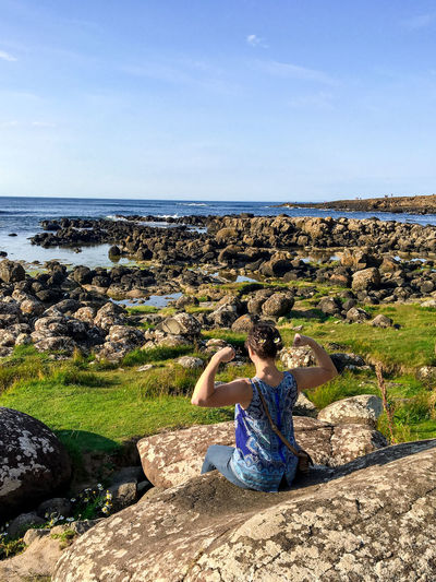 Woman flexing muscles while sitting on rock by sea against sky