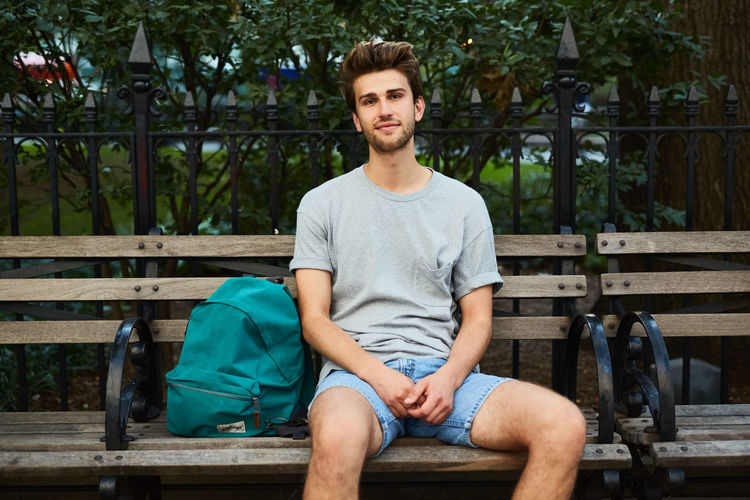 Portrait of young man sitting on bench by fence