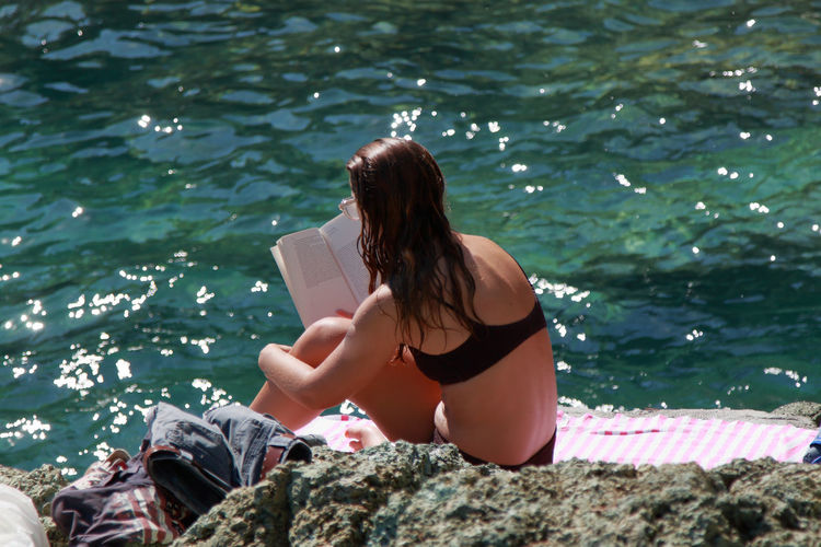 A caucasian woman reads a book sitting on a cliff by the sea